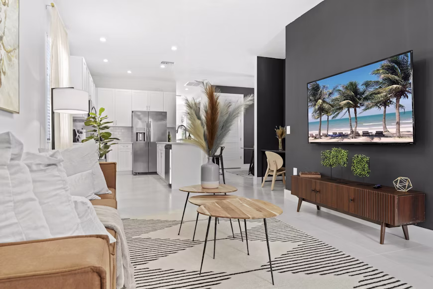 Interior de sala integrada à cozinha em conceito aberto de um apartamento moderno com sofá caramelo, mesas de centro de madeira e TV na parede cinza exibindo uma praia, ilustrando o bloco Airbnb para quem quer se sentir em casa.