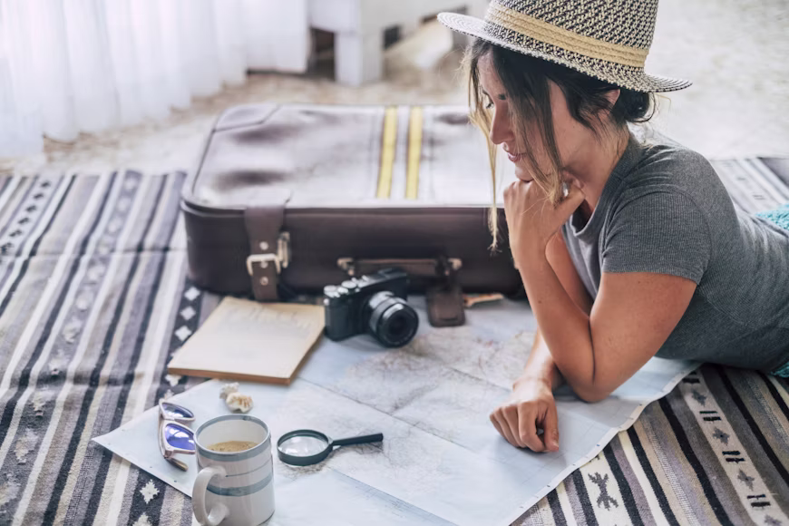 Mulher com chapéu de palha deitada no chão analisando detalhadamente um mapa com uma lupa ao lado de uma mala e câmera fotográfica, ilustrando o erro de não verificar o itinerário completo ao comprar um pacote de viagem.