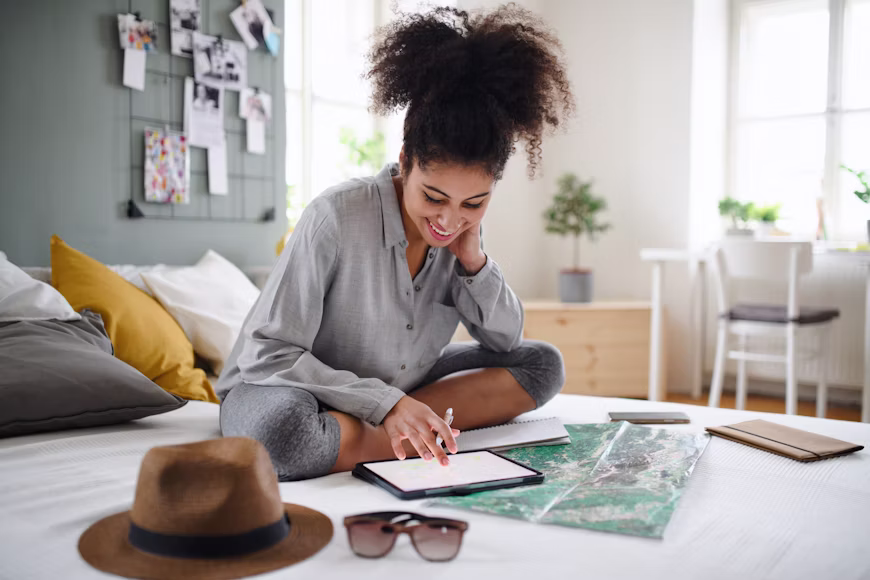 Mulher jovem sorridente sentada na cama usando tablet para desenhar seu roteiro de viagem, com mapa, chapéu e óculos de sol à sua frente, ilustrando o bloco sobre flexibilidade de horários e roteiros.
