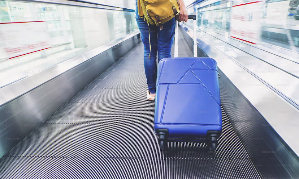 Plano focado nas pernas de uma viajante caminhando em uma esteira rolante de aeroporto, puxando uma mala rígida azul e usando mochila amarela, ilustrando o fluxo de para onde vai a mala despachada se o voo tem conexão.