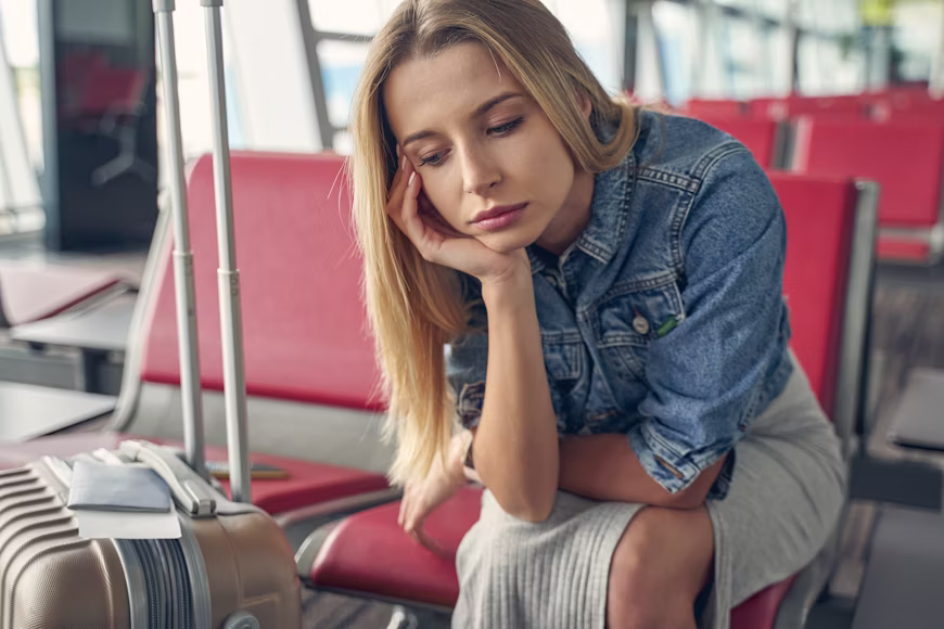 Mulher loira de jaqueta jeans com expressão de desânimo e decepção sentada em banco de aeroporto com sua mala e passaporte ao lado, ilustrando erros que fazem os viajantes gastarem mais.