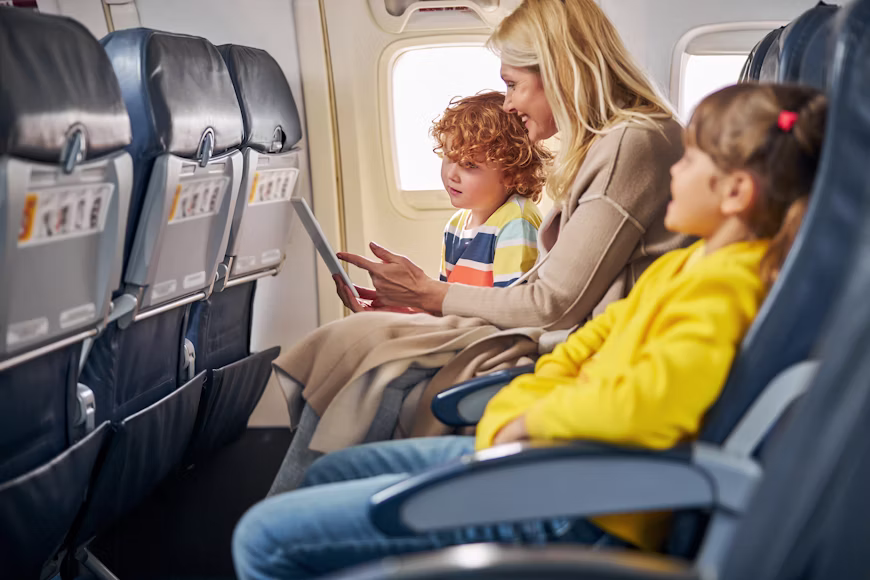 Mãe e duas crianças sentadas sorrindo na fileira de assentos de um avião comercial usando um tablet perto da janela, ilustrando as dicas sobre assento no avião como escolher o melhor lugar sem pagar taxas.