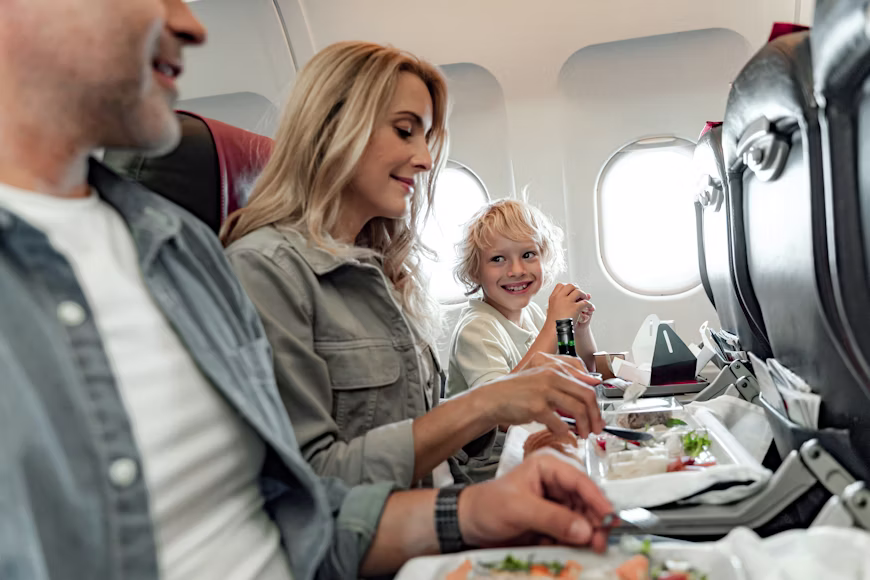 Pai, mãe e filho sentados juntos na mesma fileira da classe econômica de um avião comendo durante o serviço de bordo, ilustrando o guia de assento no avião como escolher o melhor lugar sem pagar taxas extras.