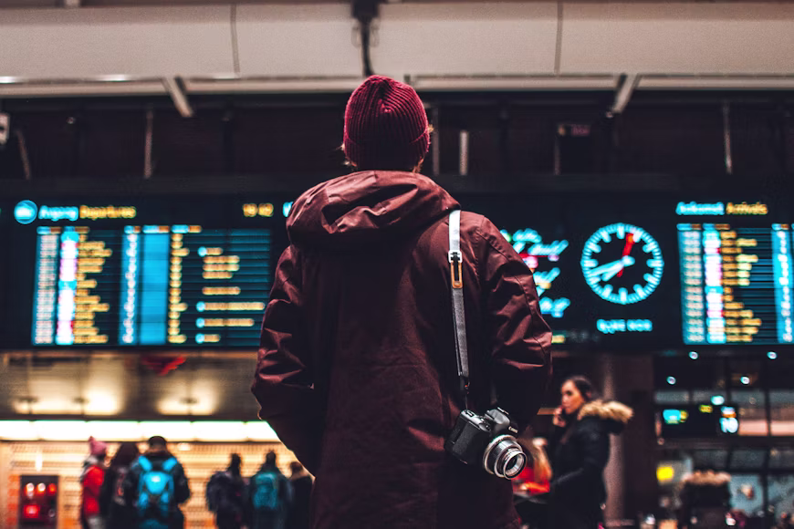 Fotógrafo de costas usando touca vermelha e jaqueta, com uma câmera fotográfica tiracolo, observando o painel de voos e um grande relógio em um aeroporto, ilustrando a análise de preços de um buscador automático de passagem aérea.