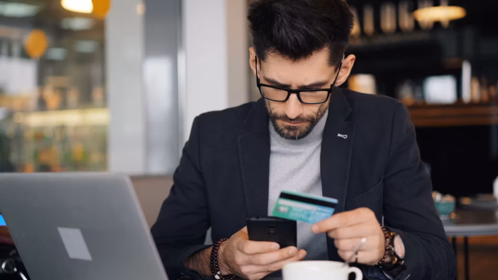Homem de óculos e paletó escuro concentrado, segurando um smartphone e um cartão de crédito em frente a um notebook em uma mesa, ilustrando táticas de como acumular milhas mais rápido.