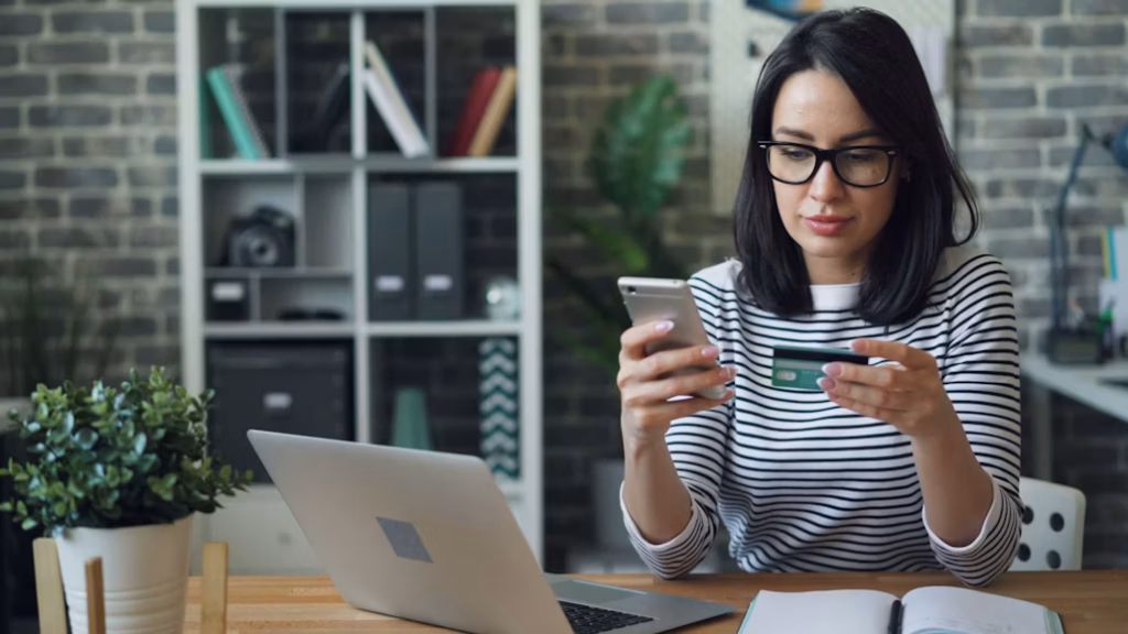 Mulher de óculos e blusa listrada sentada em uma mesa de escritório, segurando um smartphone e um cartão de crédito verde em frente ao notebook, ilustrando as estratégias de como acumular milhas no cartão de crédito.