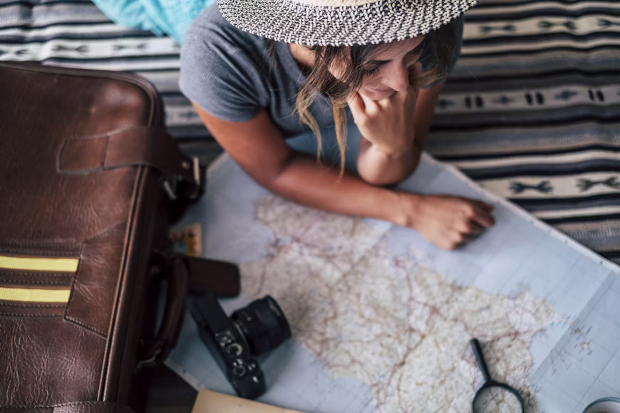 Vista superior de uma mulher de chapéu deitada olhando para um mapa aberto no chão ao lado de uma mala de couro e câmera fotográfica, ilustrando a revisão de planejamento para evitar erros ao calcular quantas milhas preciso para viajar.