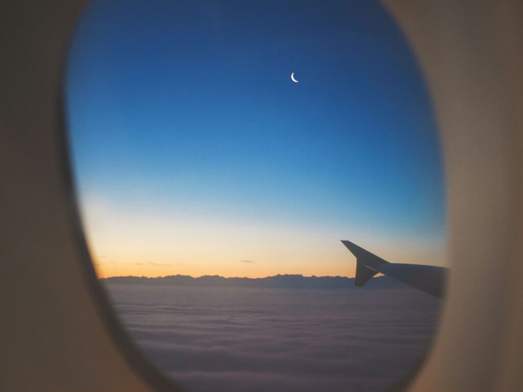 Vista através da janela de um avião mostrando a ponta da asa sobre um mar de nuvens durante o pôr do sol, com o céu azul e a lua crescente ao fundo, representando os benefícios de escolher o melhor programa de milhas.