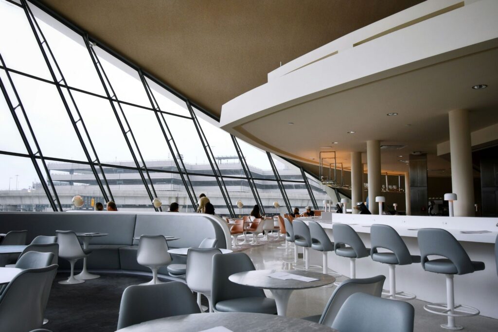 Interior de um lounge ou sala VIP de aeroporto com design moderno, mesas redondas, cadeiras cinzas e uma grande parede de vidro inclinada, ilustrando as facilidades disponíveis em conexões longas no aeroporto.