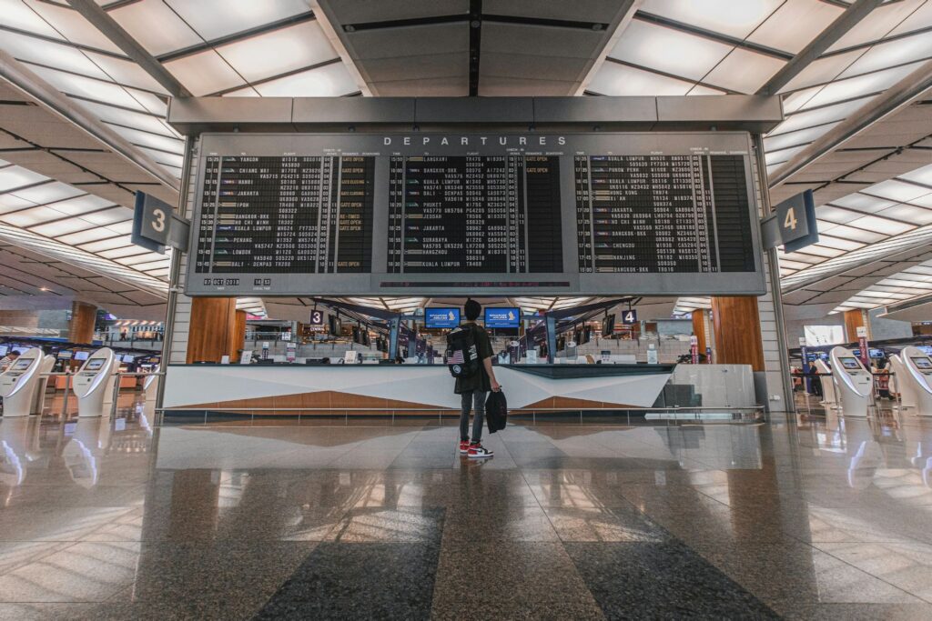 Viajante de mochila de costas parado no centro de um grande saguão de aeroporto olhando para um enorme painel de voos, ilustrando o guia de como planejar conexões longas gastando menos.