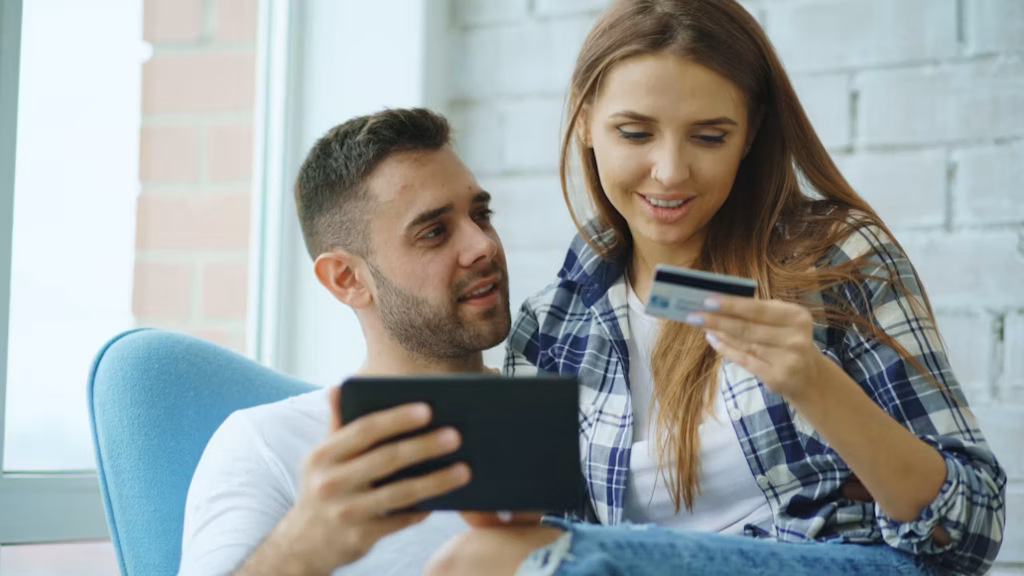 Casal sentado junto sorrindo enquanto o homem segura um tablet e a mulher analisa um cartão de crédito, ilustrando a escolha de quais programas ajudam no objetivo de como acumular milhas.