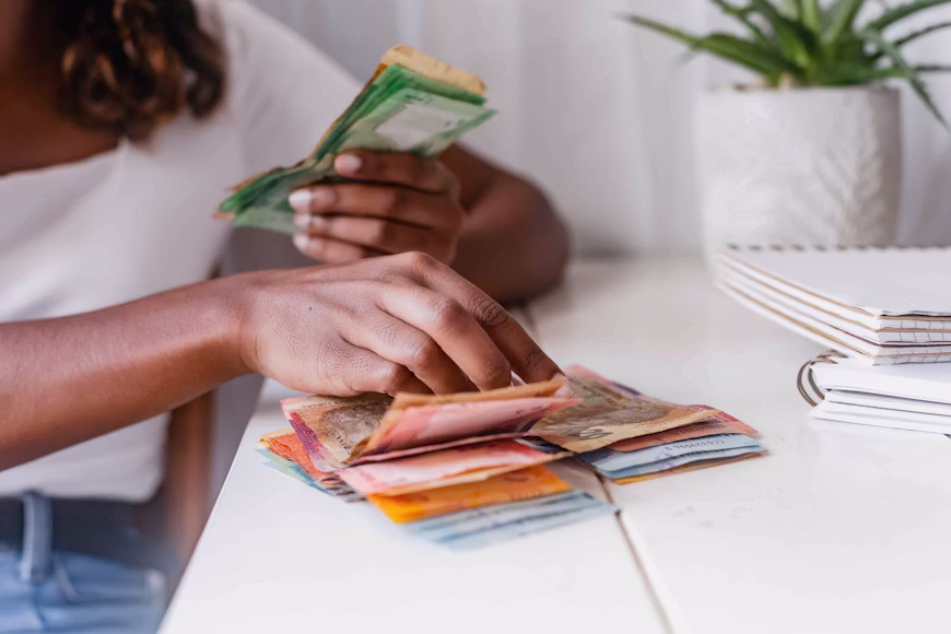 Mãos de uma pessoa organizando e contando cédulas de dinheiro coloridas sobre uma mesa branca de trabalho ao lado de cadernos, ilustrando o controle financeiro de quem busca como ganhar dinheiro com milhas.