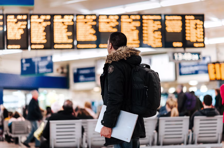 de costas usando casaco e mochila, segurando um notebook e observando o painel eletrônico de partidas e chegadas de voos no saguão do aeroporto.