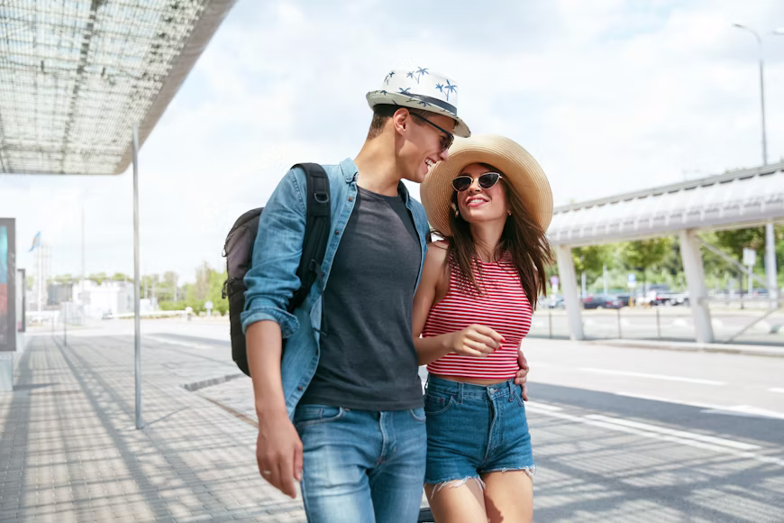 Homem com mochila e camisa jeans e mulher de blusa listrada e chapéu sorrindo e caminhando juntos na área externa de um aeroporto, ilustrando a meta alcançada de quantas milhas preciso para viajar.