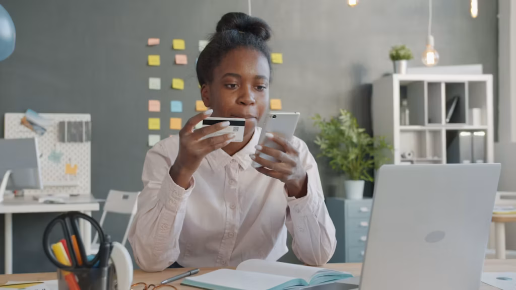 Mulher negra com cabelo preso em coque, sentada em uma mesa de trabalho, segurando um cartão com tarja magnética e olhando concentrada para o celular, ilustrando o processo de como descobrir se minhas milhas expiraram.